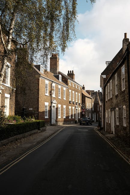 A narrow residential street with closely packed brick townhouses on both sides, some with white-framed windows and chimneys, during daytime with partly cloudy sky. A large, leafy tree overhangs the st