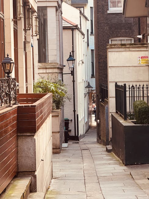 A narrow residential street with closely packed brick townhouses on both sides, some with white-framed windows and chimneys, during daytime with partly cloudy sky. A large, leafy tree overhangs the st