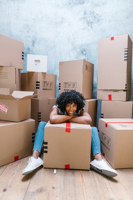 A male mover wearing a blue hoodie and dark trousers is seated on the edge of an open van or moving truck, surrounded by cardboard boxes and packing materials. He is holding and examining a document o