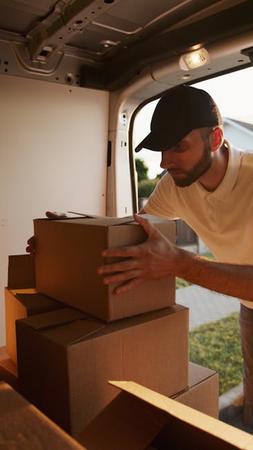 A man wearing a black cap and a beige t-shirt is inside a van, carefully placing a brown cardboard box onto a stack of similar boxes during a home relocation process. The van's interior ceiling and side panel are visible, with soft lighting illuminating the workspace. Outside the open van door, a blurred outdoor scene with greenery can be seen, indicating daytime. The boxes are securely packed with some covered in plastic wrap, and the man appears to be engaged in a packing and moving activity associated with furniture transport and relocation services. This scene exemplifies the loading process during a professional removals service provided by Man and Van Finchley, focusing on efficient and careful handling of household items.