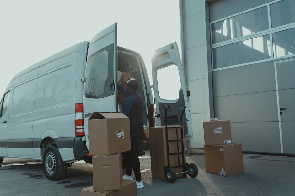 A man wearing a black cap and a beige t-shirt is inside a van, carefully placing a brown cardboard box onto a stack of similar boxes during a home relocation process. The van's interior ceiling and side panel are visible, with soft lighting illuminating the workspace. Outside the open van door, a blurred outdoor scene with greenery can be seen, indicating daytime. The boxes are securely packed with some covered in plastic wrap, and the man appears to be engaged in a packing and moving activity associated with furniture transport and relocation services. This scene exemplifies the loading process during a professional removals service provided by Man and Van Finchley, focusing on efficient and careful handling of household items.
