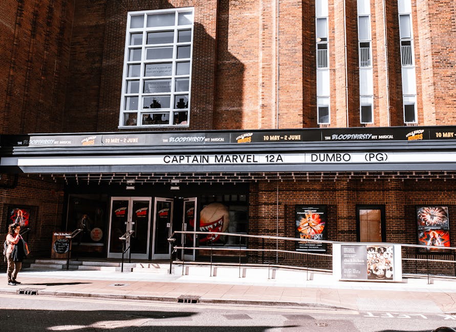 Photograph of the exterior of a red brick theatre building with large multi-pane windows and a marquee advertising the musical 'Blood Brothers' running from May 10 to June 2. Below the marquee, there are three black-framed glass doors with vertical metal handles, and to the right, a brown wooden panel is visible. In front of the building, a man and a woman are walking on the pavement; the man is carrying a bag, and the woman is holding a phone. The pavement features a tactile paving strip near the curb. Adjacent to the building, there is a metal railing with a horizontal bar, implying an accessible ramp for wheelchair access. The scene is brightly lit with direct sunlight, casting shadows on the pavement and building facade. The context relates to house removals or furniture transport involving careful handling of items during a home relocation, with [COMPANY_NAME] potentially involved in such moving services near Finchley, as indicated by the 'Removals near Phoenix Cinema Finchley access tips' page.