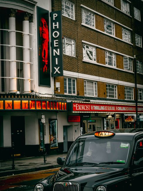 Photograph of the exterior of a red brick theatre building with large multi-pane windows and a marquee advertising the musical 'Blood Brothers' running from May 10 to June 2. Below the marquee, there are three black-framed glass doors with vertical metal handles, and to the right, a brown wooden panel is visible. In front of the building, a man and a woman are walking on the pavement; the man is carrying a bag, and the woman is holding a phone. The pavement features a tactile paving strip near the curb. Adjacent to the building, there is a metal railing with a horizontal bar, implying an accessible ramp for wheelchair access. The scene is brightly lit with direct sunlight, casting shadows on the pavement and building facade. The context relates to house removals or furniture transport involving careful handling of items during a home relocation, with [COMPANY_NAME] potentially involved in such moving services near Finchley, as indicated by the 'Removals near Phoenix Cinema Finchley access tips' page.